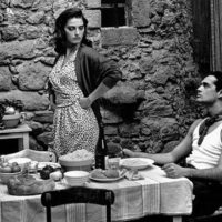 Black-and-white film still of a woman standing beside a rustic meal